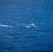 An aerial or elevated view of open blue ocean showing two vessels in proximity to each other. One is a larger white multi-deck vessel, likely a patrol or passenger boat, while the other is a smaller, faster rigid inflatable boat (RIB) leaving a prominent white wake. The scene suggests a possible intercept, escort, or patrol operation at sea.