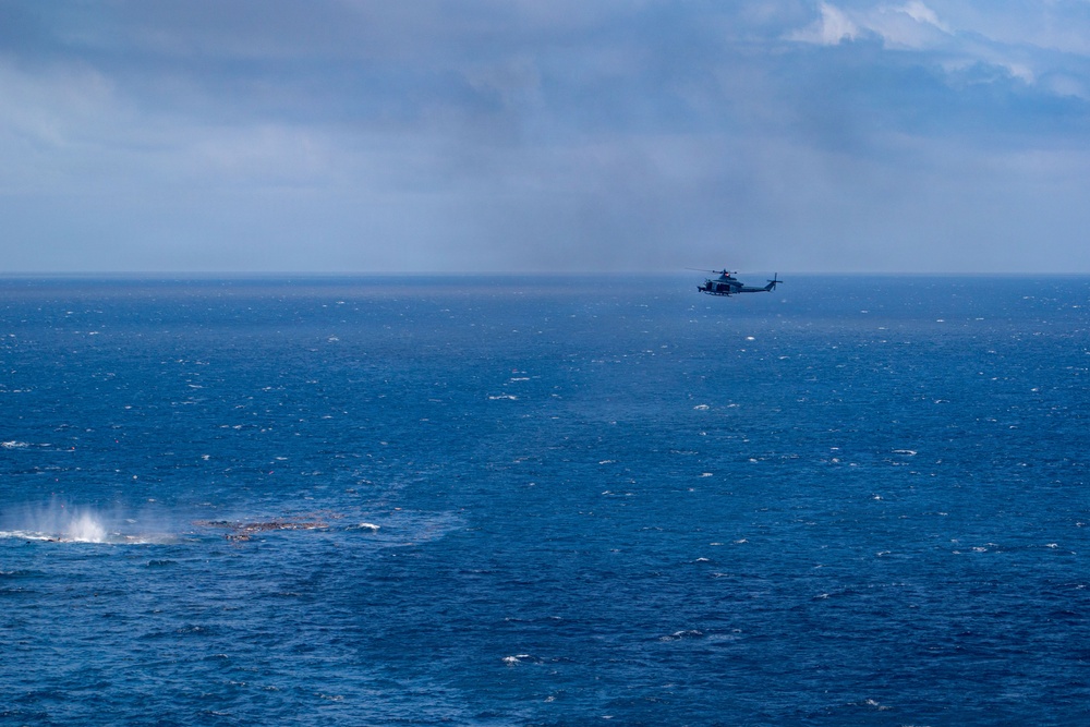 A military or coast guard helicopter flies over a choppy, deep-blue open ocean under an overcast sky. In the lower-left portion of the frame, there is a visible disturbance on the water surface, possibly a submerged reef, debris, or the aftermath of an impact or explosion. The scene suggests a maritime patrol, search-and-rescue, or surveillance operation.