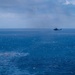 A military or coast guard helicopter flies over a choppy, deep-blue open ocean under an overcast sky. In the lower-left portion of the frame, there is a visible disturbance on the water surface, possibly a submerged reef, debris, or the aftermath of an impact or explosion. The scene suggests a maritime patrol, search-and-rescue, or surveillance operation.