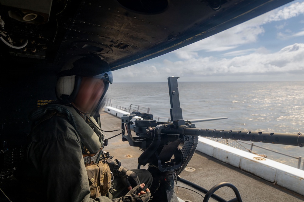 A military personnel wearing a flight helmet and olive drab flight suit is positioned at a door gun mount aboard what appears to be a military helicopter on a ship's deck. The gunner is manning a belt-fed machine gun (consistent with an M240 or similar weapon) pointed outward over open ocean. The scene suggests a naval aviation operation or shipboard security exercise.