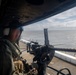 A military personnel wearing a flight helmet and olive drab flight suit is positioned at a door gun mount aboard what appears to be a military helicopter on a ship's deck. The gunner is manning a belt-fed machine gun (consistent with an M240 or similar weapon) pointed outward over open ocean. The scene suggests a naval aviation operation or shipboard security exercise.