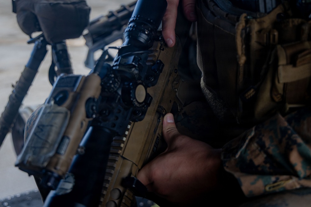 A close-up image showing a military service member in woodland MARPAT camouflage uniform handling a tan/flat dark earth colored rifle with optical scope and accessories. The weapon appears to be an M27 IAR or similar HK416-pattern rifle with a magnified optic mounted on top. A bipod-equipped weapon is also visible in the background.