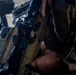 A close-up image showing a military service member in woodland MARPAT camouflage uniform handling a tan/flat dark earth colored rifle with optical scope and accessories. The weapon appears to be an M27 IAR or similar HK416-pattern rifle with a magnified optic mounted on top. A bipod-equipped weapon is also visible in the background.