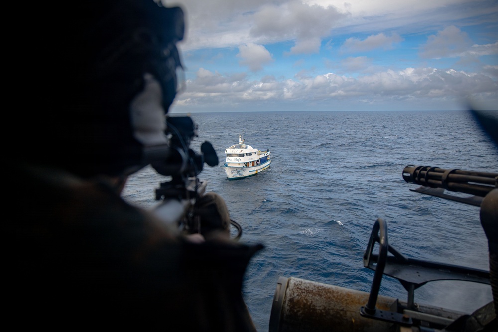 An aerial or elevated view from a military or law enforcement helicopter showing an armed operator aiming a weapon toward a white civilian-style vessel on open ocean. The foreground shows a helmeted figure with a scoped weapon and what appears to be a multi-barrel rotary cannon (minigun) mounted on the aircraft. The scene suggests a maritime interdiction or surveillance operation.