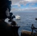An aerial or elevated view from a military or law enforcement helicopter showing an armed operator aiming a weapon toward a white civilian-style vessel on open ocean. The foreground shows a helmeted figure with a scoped weapon and what appears to be a multi-barrel rotary cannon (minigun) mounted on the aircraft. The scene suggests a maritime interdiction or surveillance operation.