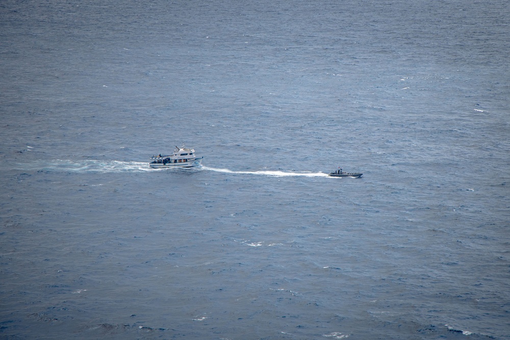 An aerial photograph shows two vessels on open water, with a larger white cabin cruiser or passenger vessel on the left and a smaller, darker rigid inflatable boat (RIB) or patrol craft on the right. Both vessels appear to be underway, moving in the same general direction with visible wake trails behind them. The image was taken from a significant altitude, suggesting it was captured from an aircraft or elevated vantage point.