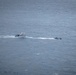 An aerial photograph shows two vessels on open water, with a larger white cabin cruiser or passenger vessel on the left and a smaller, darker rigid inflatable boat (RIB) or patrol craft on the right. Both vessels appear to be underway, moving in the same general direction with visible wake trails behind them. The image was taken from a significant altitude, suggesting it was captured from an aircraft or elevated vantage point.