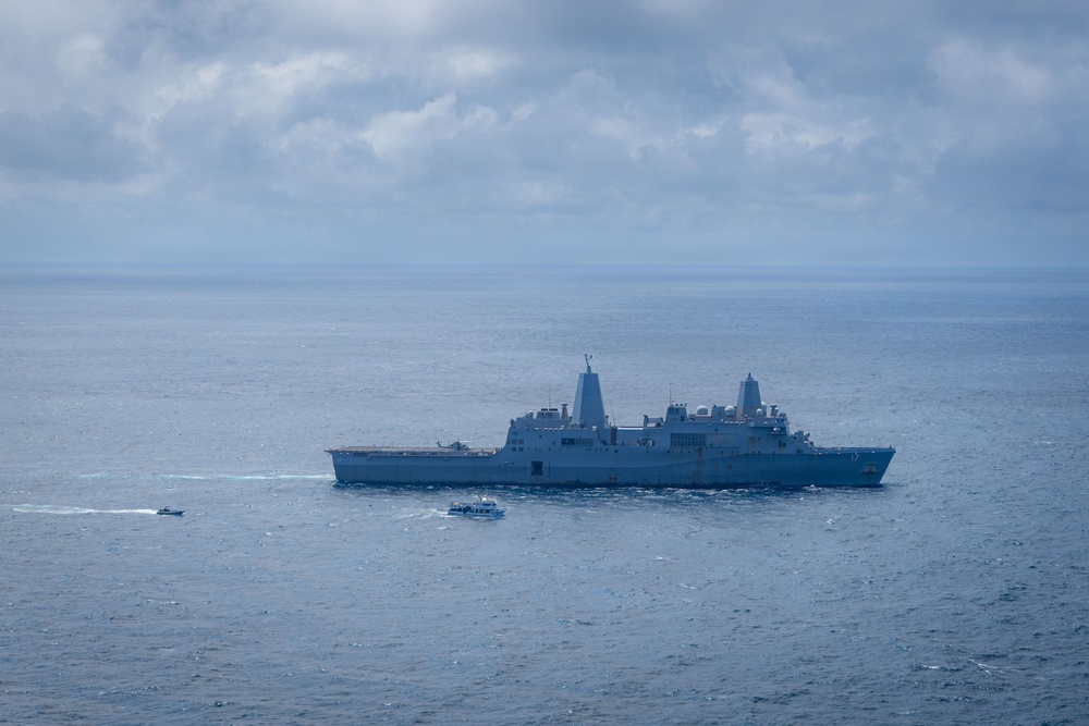 A large gray naval warship, consistent with a US Navy amphibious transport dock (LPD-class), is underway on open ocean under overcast skies. Two smaller civilian or patrol vessels are visible in the foreground, significantly smaller in scale compared to the warship. The scene appears to be a maritime operation or transit in open blue-gray waters.