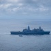 A large gray naval warship, consistent with a US Navy amphibious transport dock (LPD-class), is underway on open ocean under overcast skies. Two smaller civilian or patrol vessels are visible in the foreground, significantly smaller in scale compared to the warship. The scene appears to be a maritime operation or transit in open blue-gray waters.
