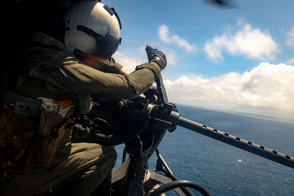 A military aircrew member wearing a flight helmet and olive drab flight suit is operating a door-mounted machine gun from an open helicopter door over open ocean. The gunner is positioned at the weapon system, which appears to be an M240 or similar belt-fed machine gun mounted on a pintle or swing arm. The image is taken from inside the aircraft looking outward, with blue sky, clouds, and ocean visible in the background.