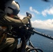 A military aircrew member wearing a flight helmet and olive drab flight suit is operating a door-mounted machine gun from an open helicopter door over open ocean. The gunner is positioned at the weapon system, which appears to be an M240 or similar belt-fed machine gun mounted on a pintle or swing arm. The image is taken from inside the aircraft looking outward, with blue sky, clouds, and ocean visible in the background.