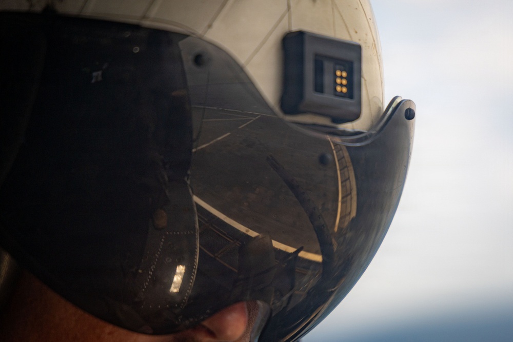 A close-up photograph of a military flight helmet with a dark tinted visor in the down position. A black electronic module with gold contact pins is mounted on the upper right side of the white helmet. The visor reflects what appears to be a large military aircraft, likely a bomber or transport plane.