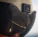 A close-up photograph of a military flight helmet with a dark tinted visor in the down position. A black electronic module with gold contact pins is mounted on the upper right side of the white helmet. The visor reflects what appears to be a large military aircraft, likely a bomber or transport plane.