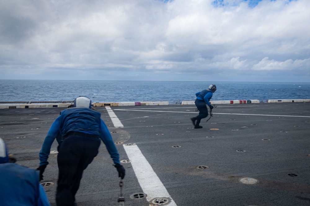 The image shows personnel in blue uniforms and helmets working on what appears to be a naval vessel's flight deck over open ocean. Two or more crew members are moving across the deck, which features white painted lines, tie-down rings, and deck markings typical of an aircraft carrier or amphibious assault ship. The scene is captured under a partly cloudy sky with calm ocean visible in the background.