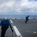The image shows personnel in blue uniforms and helmets working on what appears to be a naval vessel's flight deck over open ocean. Two or more crew members are moving across the deck, which features white painted lines, tie-down rings, and deck markings typical of an aircraft carrier or amphibious assault ship. The scene is captured under a partly cloudy sky with calm ocean visible in the background.