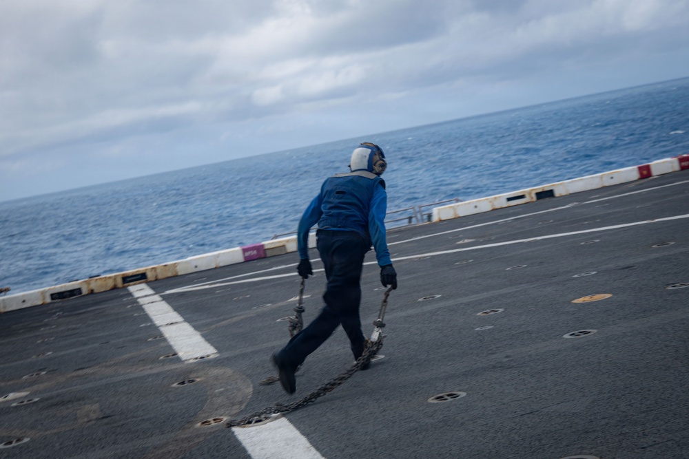 A naval deck crew member dressed in blue coveralls is running across what appears to be an aircraft carrier flight deck while carrying metal chains. The person is wearing hearing protection and gloves, standard safety gear for flight deck operations. The ocean is visible in the background under an overcast sky.