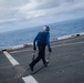 A naval deck crew member dressed in blue coveralls is running across what appears to be an aircraft carrier flight deck while carrying metal chains. The person is wearing hearing protection and gloves, standard safety gear for flight deck operations. The ocean is visible in the background under an overcast sky.