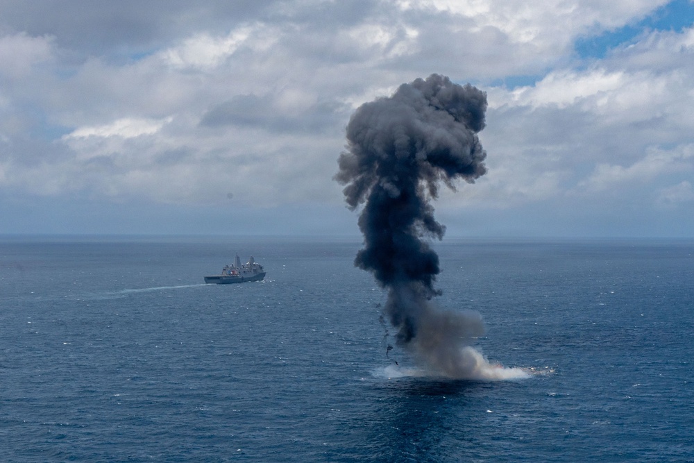 A large explosion or detonation is occurring on the open ocean surface, producing a tall column of dark black smoke rising into a partly cloudy sky. A naval vessel is visible in the background to the left, suggesting this may be a military exercise, weapons test, or controlled sinking (SINKEX) operation. The explosion appears to originate at the waterline, with debris and white spray visible at the base.