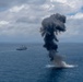 A large explosion or detonation is occurring on the open ocean surface, producing a tall column of dark black smoke rising into a partly cloudy sky. A naval vessel is visible in the background to the left, suggesting this may be a military exercise, weapons test, or controlled sinking (SINKEX) operation. The explosion appears to originate at the waterline, with debris and white spray visible at the base.