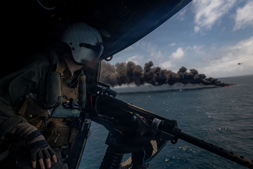 A military helicopter door gunner is positioned at the open door of an aircraft, manning a machine gun while observing a vessel on fire in the ocean below. The scene depicts what appears to be a maritime interdiction or combat operation, with a burning boat producing a large plume of dark smoke on the water's surface. A second helicopter is faintly visible in the background on the right side of the frame.