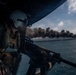 A military helicopter door gunner is positioned at the open door of an aircraft, manning a machine gun while observing a vessel on fire in the ocean below. The scene depicts what appears to be a maritime interdiction or combat operation, with a burning boat producing a large plume of dark smoke on the water's surface. A second helicopter is faintly visible in the background on the right side of the frame.