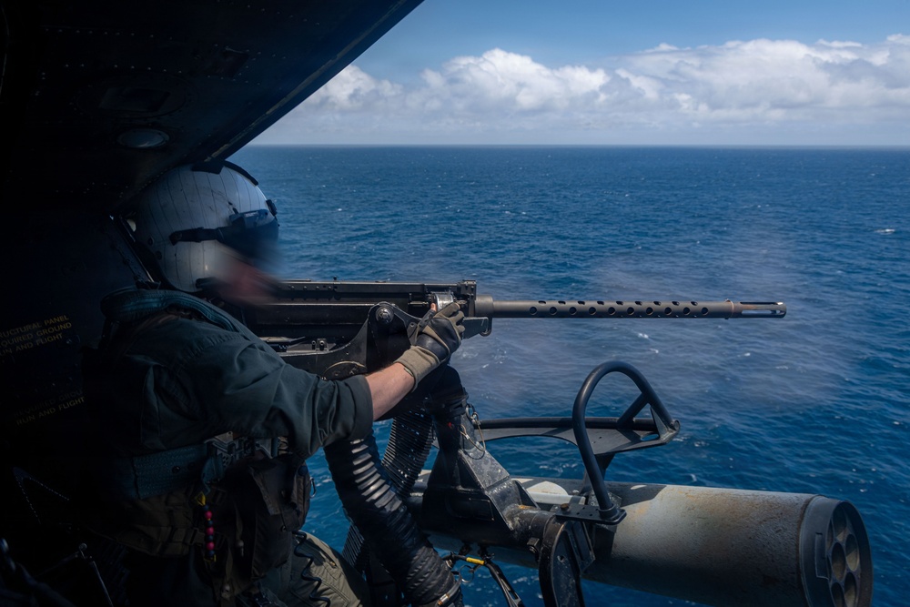 A military aircrew member is positioned at the open door of a helicopter over open ocean, operating a mounted machine gun. The operator is wearing full flight gear including a helmet and flight suit. The image appears to depict a live-fire or readiness exercise over a blue ocean environment.