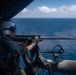 A military aircrew member is positioned at the open door of a helicopter over open ocean, operating a mounted machine gun. The operator is wearing full flight gear including a helmet and flight suit. The image appears to depict a live-fire or readiness exercise over a blue ocean environment.