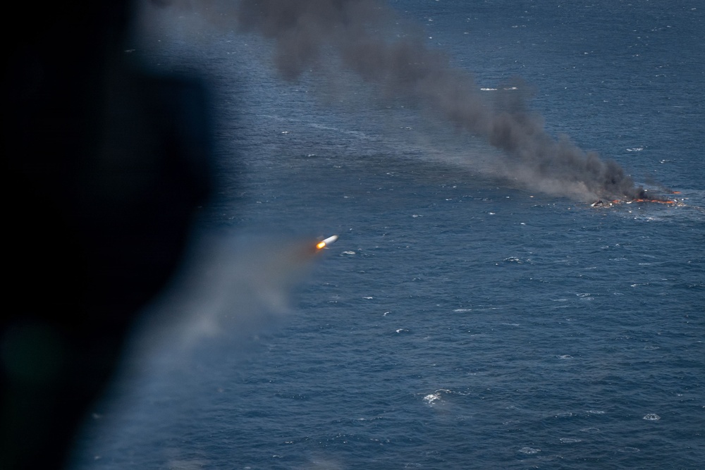 An aerial photograph taken from an aircraft shows a missile or rocket in flight over open ocean, with a burning target or vessel visible in the background producing thick black smoke. The image appears to document a live-fire military exercise or weapons test conducted at sea. The projectile is clearly visible with an active propulsion flame trailing behind it.