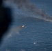 An aerial photograph taken from an aircraft shows a missile or rocket in flight over open ocean, with a burning target or vessel visible in the background producing thick black smoke. The image appears to document a live-fire military exercise or weapons test conducted at sea. The projectile is clearly visible with an active propulsion flame trailing behind it.