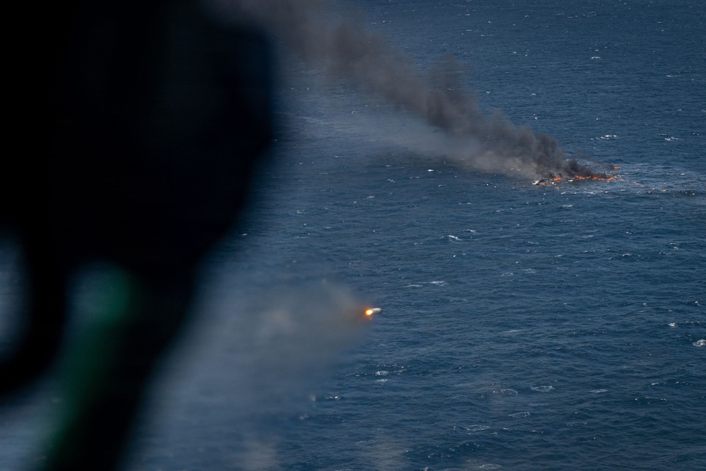 An aerial photograph taken from inside an aircraft showing a burning object on the ocean surface, producing a large column of dark smoke. A second bright point of light, possibly a flare or projectile, is visible closer to the camera in the lower center of the frame. The image appears to document a maritime incident, possibly a military exercise, vessel destruction, or emergency situation at sea.