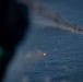 An aerial photograph taken from inside an aircraft showing a burning object on the ocean surface, producing a large column of dark smoke. A second bright point of light, possibly a flare or projectile, is visible closer to the camera in the lower center of the frame. The image appears to document a maritime incident, possibly a military exercise, vessel destruction, or emergency situation at sea.