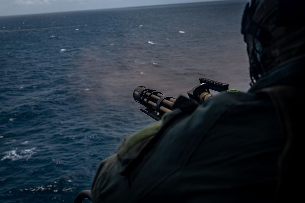 A military personnel is firing a multi-barrel rotary machine gun (consistent with an M134 Minigun) from what appears to be a helicopter or aircraft over open ocean. Smoke or gas discharge is visible near the muzzle, indicating active firing. The shooter is wearing tactical gear including a helmet and body armor.