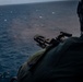 A military personnel is firing a multi-barrel rotary machine gun (consistent with an M134 Minigun) from what appears to be a helicopter or aircraft over open ocean. Smoke or gas discharge is visible near the muzzle, indicating active firing. The shooter is wearing tactical gear including a helmet and body armor.