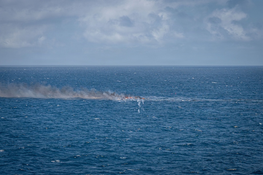The image shows an open ocean scene with smoke and what appears to be fire or burning debris visible at the water's surface at mid-distance. Dark gray smoke rises and drifts horizontally, suggesting wind conditions. The source appears to be a low-profile burning object or vessel at the waterline.