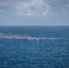 The image shows an open ocean scene with smoke and what appears to be fire or burning debris visible at the water's surface at mid-distance. Dark gray smoke rises and drifts horizontally, suggesting wind conditions. The source appears to be a low-profile burning object or vessel at the waterline.