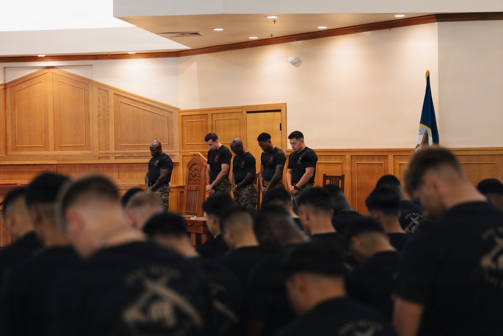A group of individuals dressed in matching black t-shirts and camouflage pants are gathered in what appears to be a military chapel or ceremonial hall. All visible persons have their heads bowed in a moment of prayer or reflection. A blue military flag is visible in the background on the right side.