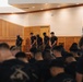 A group of individuals dressed in matching black t-shirts and camouflage pants are gathered in what appears to be a military chapel or ceremonial hall. All visible persons have their heads bowed in a moment of prayer or reflection. A blue military flag is visible in the background on the right side.