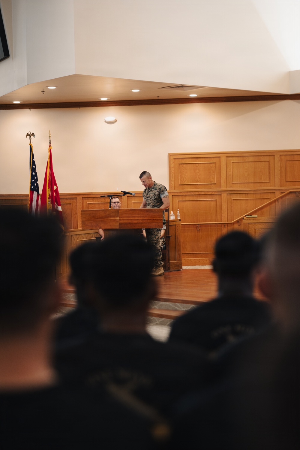 A military service member in camouflage uniform stands at a wooden podium addressing an audience in what appears to be a formal military briefing or ceremony hall. Two flags are visible in the background — an American flag and what appears to be a Marine Corps flag. A second individual is seated behind the podium area, and the foreground shows the backs of heads of seated attendees.
