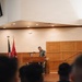 A military service member in camouflage uniform stands at a wooden podium addressing an audience in what appears to be a formal military briefing or ceremony hall. Two flags are visible in the background — an American flag and what appears to be a Marine Corps flag. A second individual is seated behind the podium area, and the foreground shows the backs of heads of seated attendees.