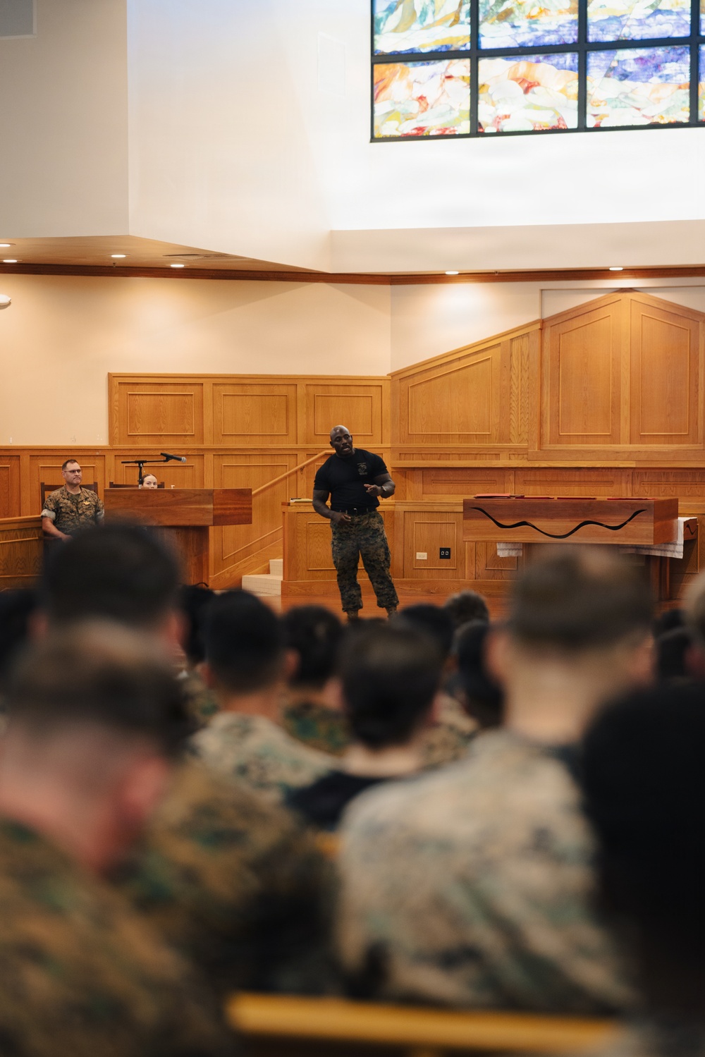 A military briefing or presentation is taking place inside a chapel or religious facility, identifiable by the stained glass window and wooden altar area. A speaker in a black shirt and military camouflage trousers stands at the front addressing a seated audience of uniformed military personnel. A second uniformed individual stands near a podium to the left of the speaker.