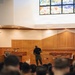 A military briefing or presentation is taking place inside a chapel or religious facility, identifiable by the stained glass window and wooden altar area. A speaker in a black shirt and military camouflage trousers stands at the front addressing a seated audience of uniformed military personnel. A second uniformed individual stands near a podium to the left of the speaker.