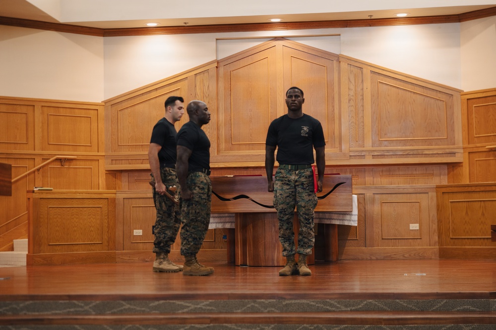 Three individuals in U.S. Marine Corps uniforms — black t-shirts and woodland MARPAT camouflage trousers with tan combat boots — are standing on a raised wooden stage inside what appears to be a military chapel or formal hall. One Marine faces forward while two others are turned slightly, suggesting a formation or rehearsal activity. The setting features warm wood paneling, recessed lighting, and a wooden altar or podium, consistent with a chapel interior.