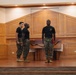 Three individuals in U.S. Marine Corps uniforms — black t-shirts and woodland MARPAT camouflage trousers with tan combat boots — are standing on a raised wooden stage inside what appears to be a military chapel or formal hall. One Marine faces forward while two others are turned slightly, suggesting a formation or rehearsal activity. The setting features warm wood paneling, recessed lighting, and a wooden altar or podium, consistent with a chapel interior.