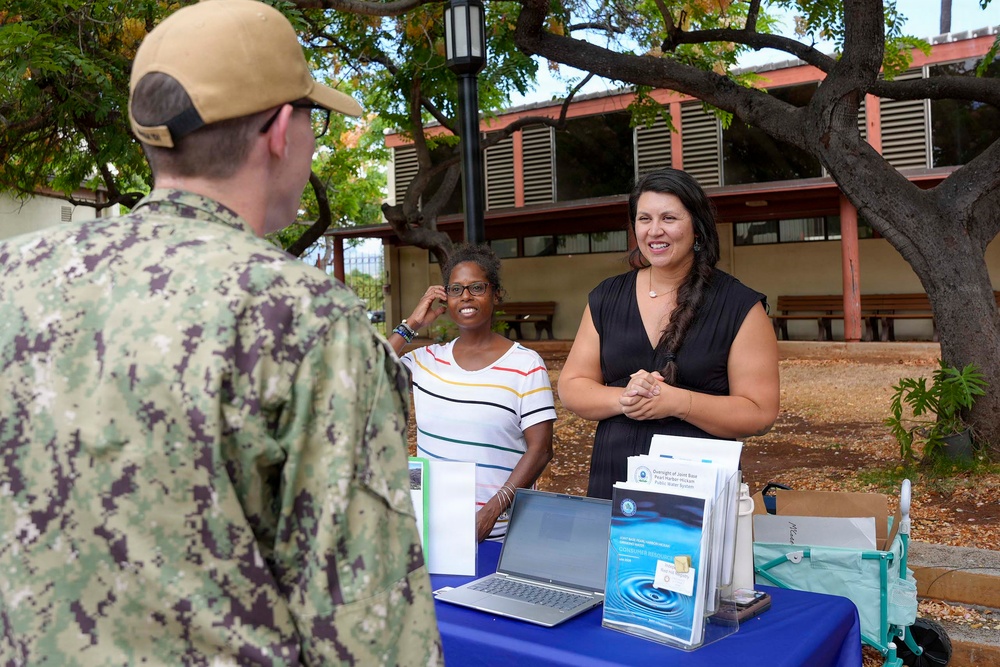 A U.S. Navy service member in digital camouflage uniform is speaking with two civilian women at an outdoor information booth. The table displays materials related to water system oversight, including a laptop, brochures, and documents referencing Pearl Harbor-Hickam. The setting appears to be a military installation in Hawaii, likely during a community outreach or informational event.