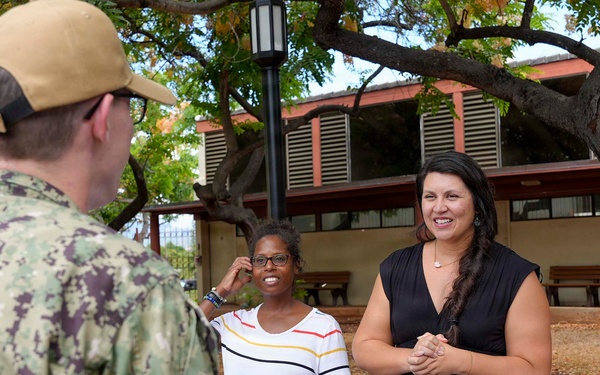 A U.S. Navy service member in digital camouflage uniform is speaking with two civilian women at an outdoor information booth. The table displays materials related to water system oversight, including a laptop, brochures, and documents referencing Pearl Harbor-Hickam. The setting appears to be a military installation in Hawaii, likely during a community outreach or informational event.