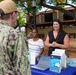A U.S. Navy service member in digital camouflage uniform is speaking with two civilian women at an outdoor information booth. The table displays materials related to water system oversight, including a laptop, brochures, and documents referencing Pearl Harbor-Hickam. The setting appears to be a military installation in Hawaii, likely during a community outreach or informational event.