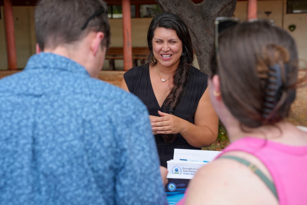 A woman in a black sleeveless top is smiling and engaged in conversation with two other individuals in an outdoor setting. She appears to be at some kind of informational or community event, as printed materials and brochures are visible. The setting appears to be a park or outdoor pavilion area.