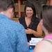 A woman in a black sleeveless top is smiling and engaged in conversation with two other individuals in an outdoor setting. She appears to be at some kind of informational or community event, as printed materials and brochures are visible. The setting appears to be a park or outdoor pavilion area.