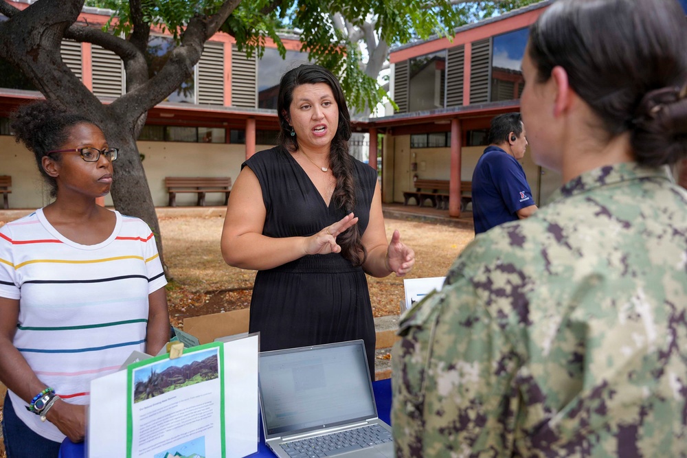 An outdoor informational or outreach event is taking place in what appears to be a courtyard or campus setting. A woman in a black dress is actively presenting or explaining something to a person in U.S. Navy digital camouflage (NWU) uniform, while another woman in a striped shirt stands nearby. A table with a laptop, printed materials, and a green folder is visible in the foreground.