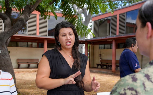 An outdoor informational or outreach event is taking place in what appears to be a courtyard or campus setting. A woman in a black dress is actively presenting or explaining something to a person in U.S. Navy digital camouflage (NWU) uniform, while another woman in a striped shirt stands nearby. A table with a laptop, printed materials, and a green folder is visible in the foreground.